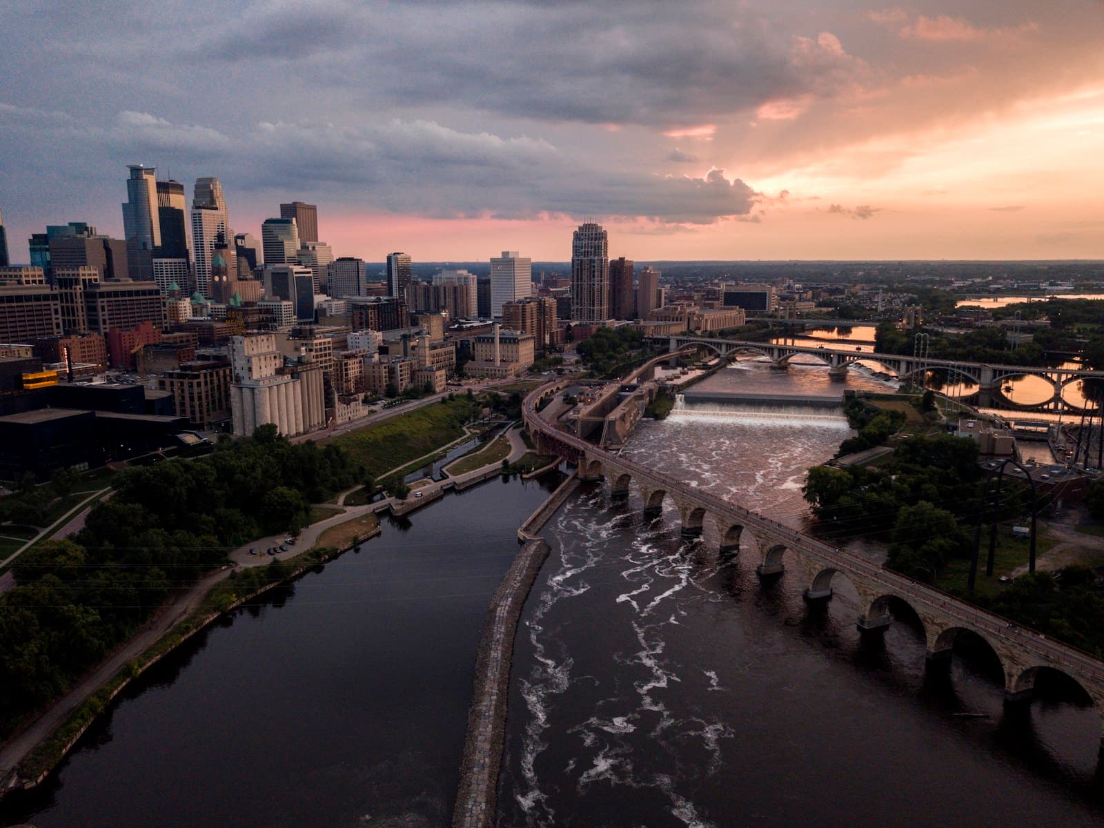 A city skyline rises beyond a stone arch bridge at sunset.